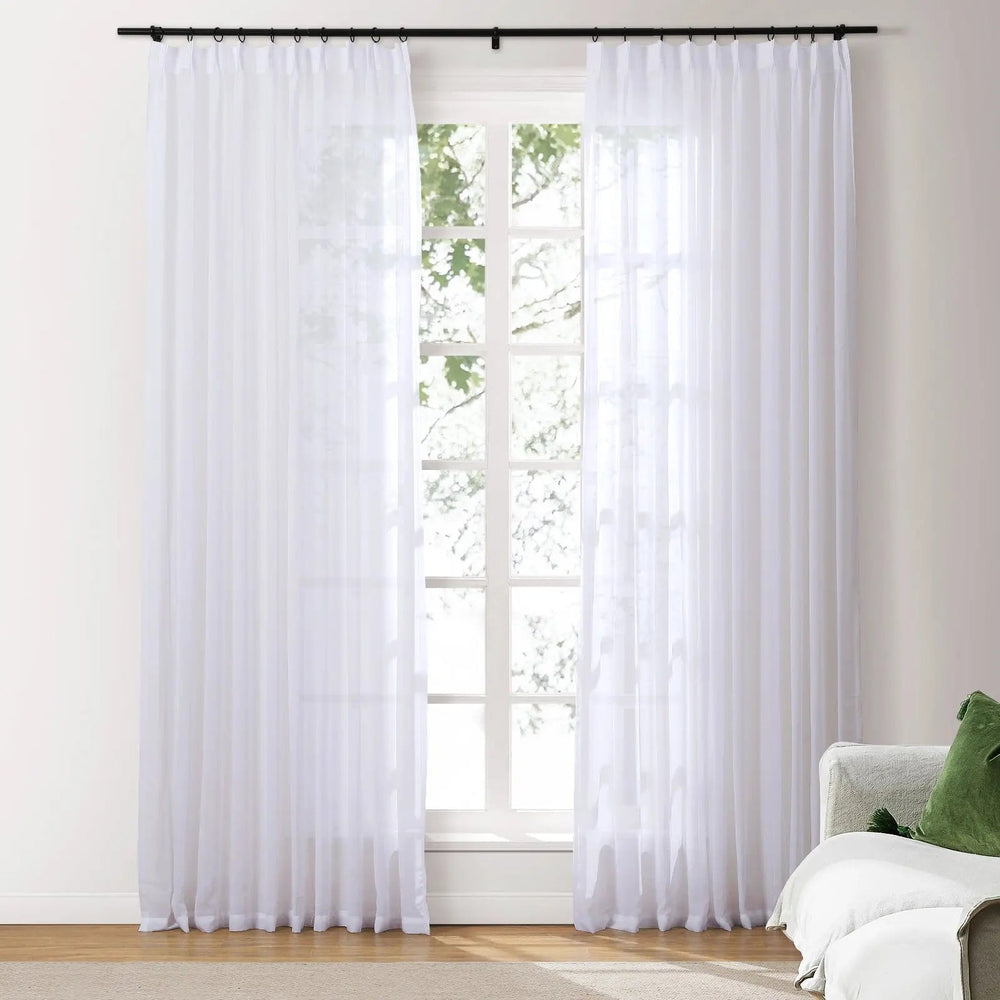 White sheer curtains hanging in front of a window with a view of greenery, in a room with a couch and green pillow.