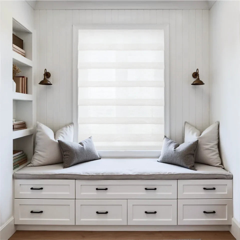 Window seat with cushions and built-in drawers in a room with white walls and wooden floor.
