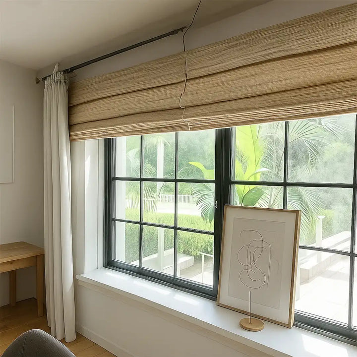 Window with wooden blinds and curtains, framed artwork on a windowsill, and a view of greenery outside.