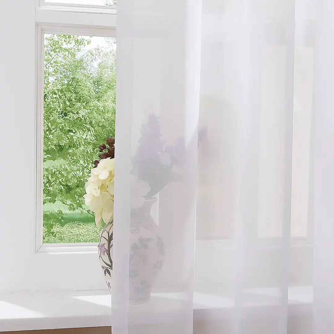 Soft, sheer white curtains draped in front of a window, with a floral vase and bouquet visible behind the fabric and greenery outside.