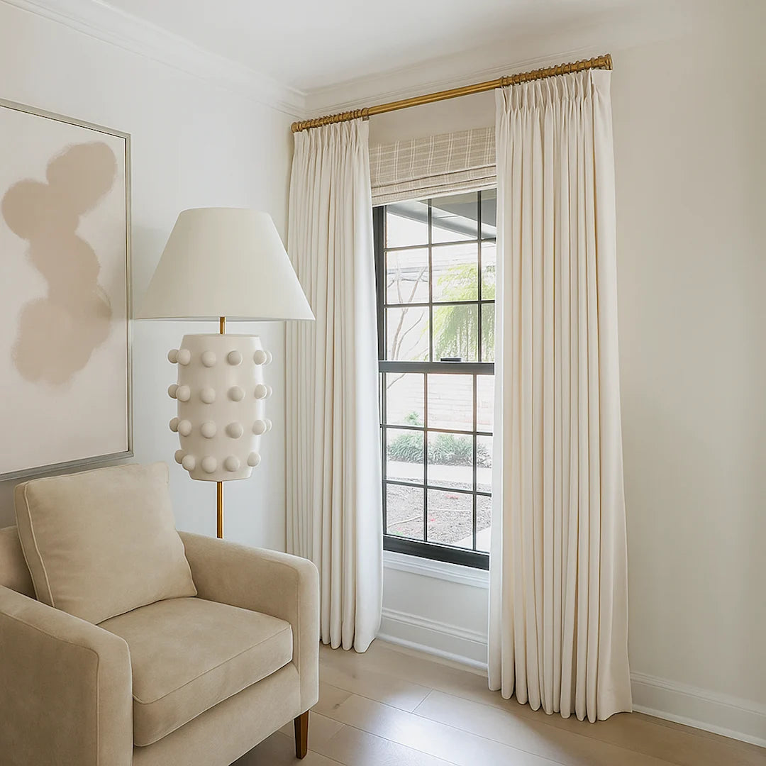 Cream-colored pleated drapes hung on a brass curtain rod beside a beige armchair and a sculptural floor lamp, framing a black-trimmed window in a bright, minimalist room.