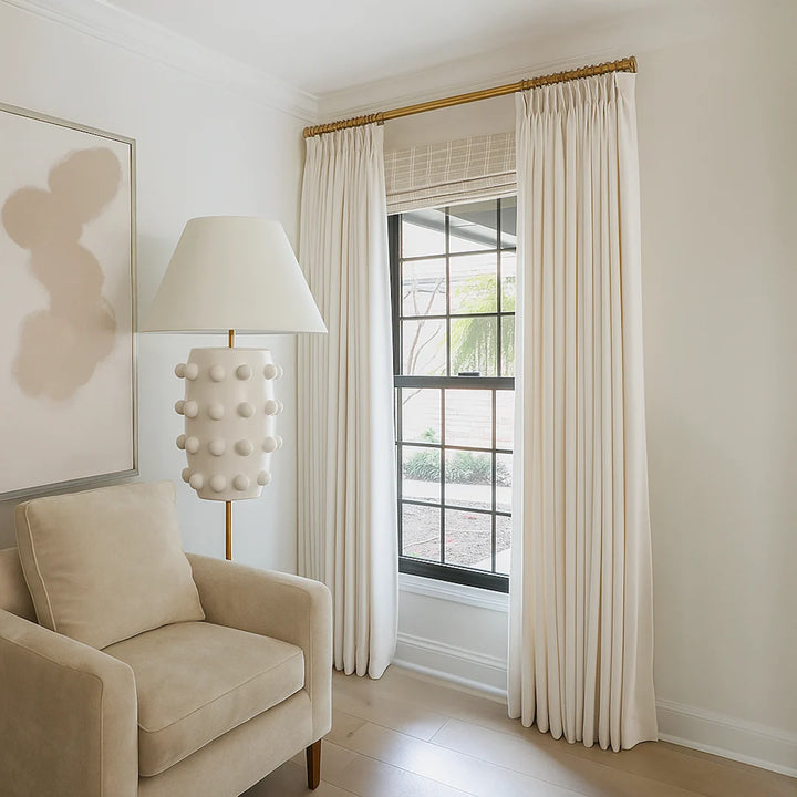 Cream-colored pleated drapes hung on a brass curtain rod beside a beige armchair and a sculptural floor lamp, framing a black-trimmed window in a bright, minimalist room.